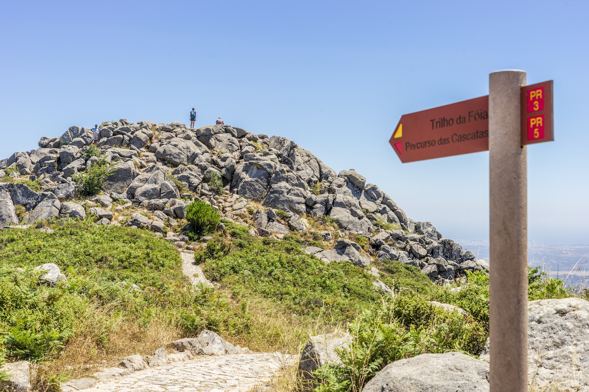A sign points in the direction of Foia, the highest point of the Algarve in Portugal. A hiker stands on top of the peak in the background.
