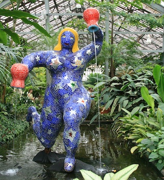 A colorful blue statue with bright yellow hair in the shape of a woman holding two orange pots with water pouring out of them. The statue is surrounded by greenery inside the Garfield Park Conservatory.  