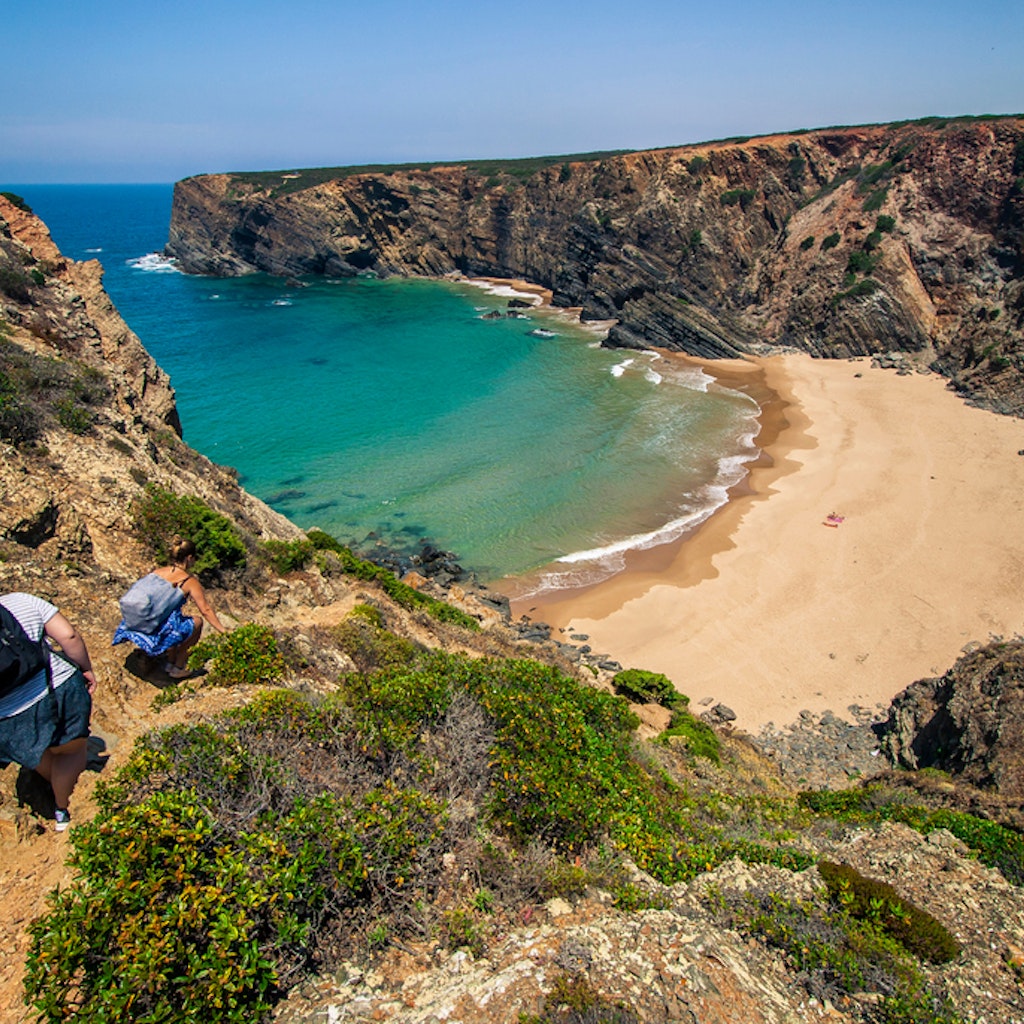 Landscape photo of two women climbing down a hill to a big, beautiful, nearly deserted beach with turquoise waters. Shot in Parque Natural do Sudoeste Alentejano e Costa Vicentina, Portugal. 