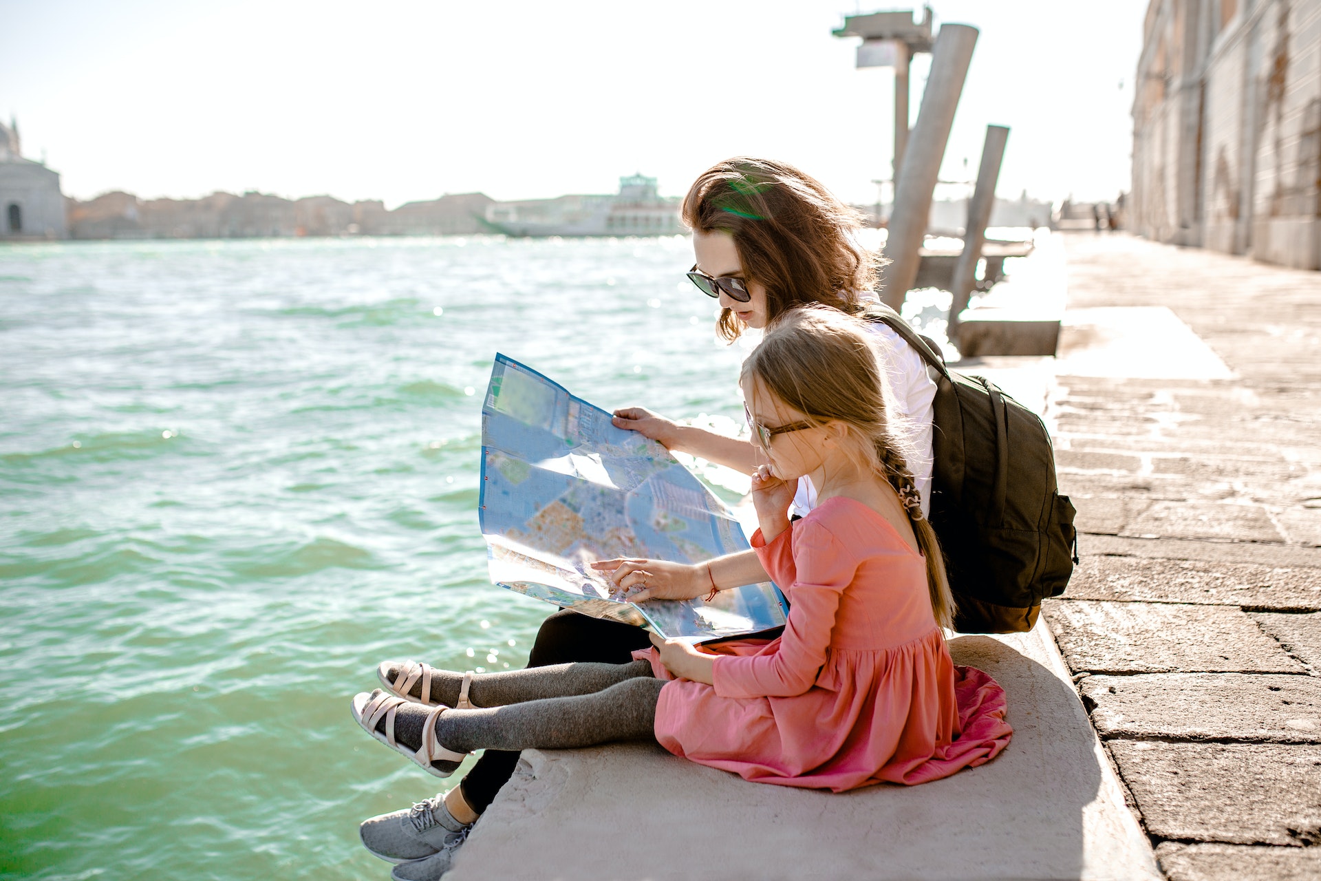 A woman and her daughter sitting on the edge of a canal in Venice as she reads a paper map