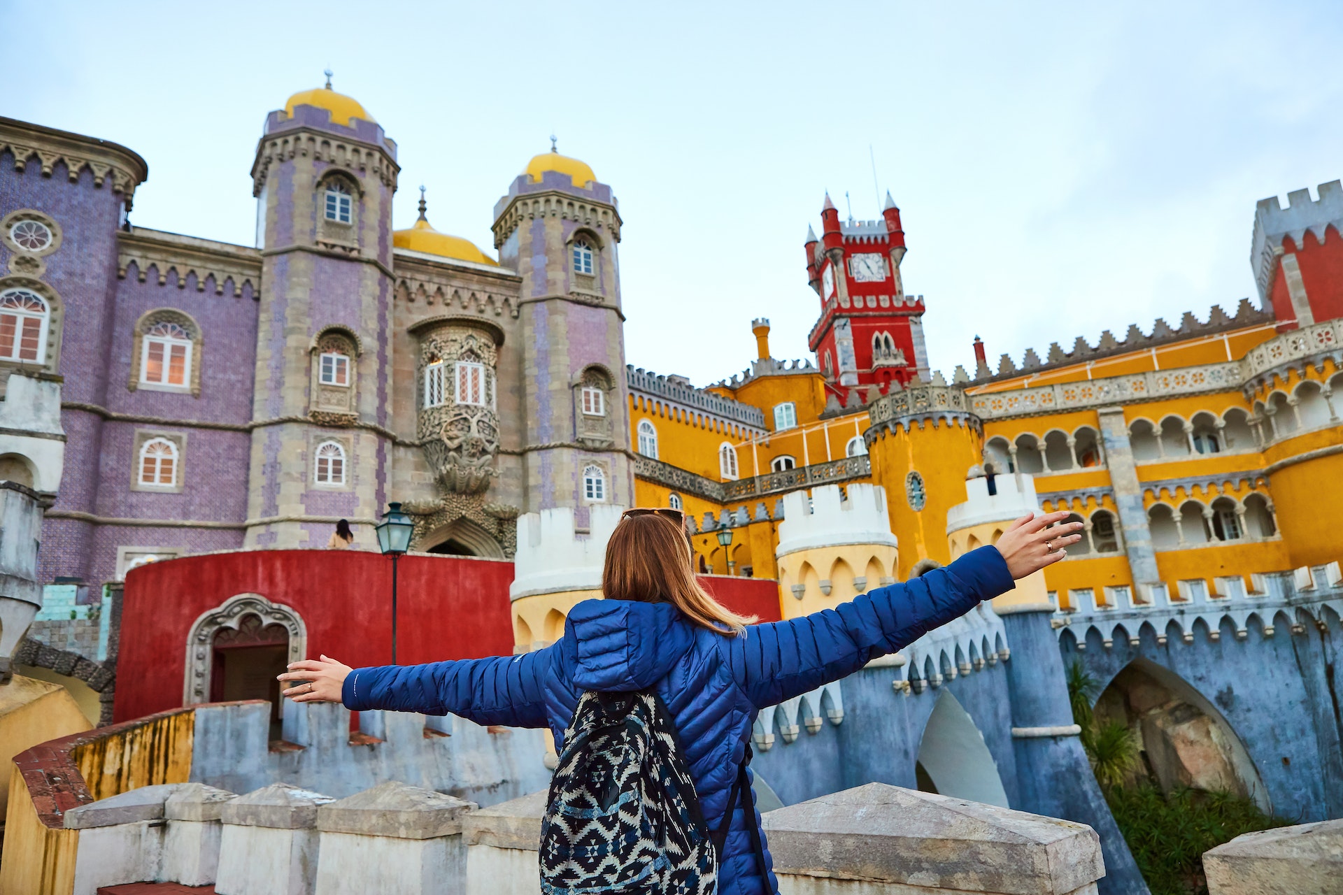 A young woman tourist opens her arms whilst looking at the colourful Pena Palace in Sintra, Portugal 