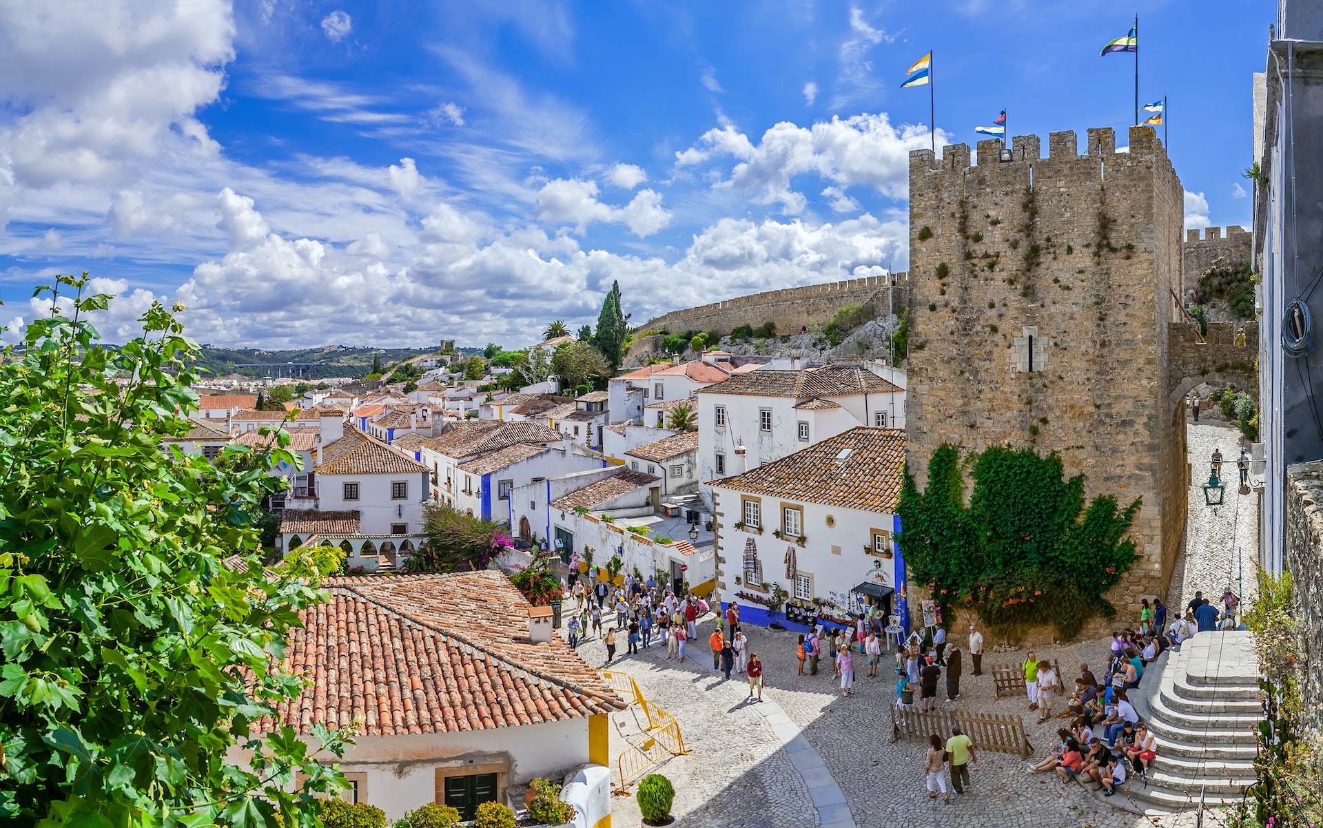 An aerial view of the medieval houses, wall and tower of Obidos, Portugal. The street is filled with crowds of people.