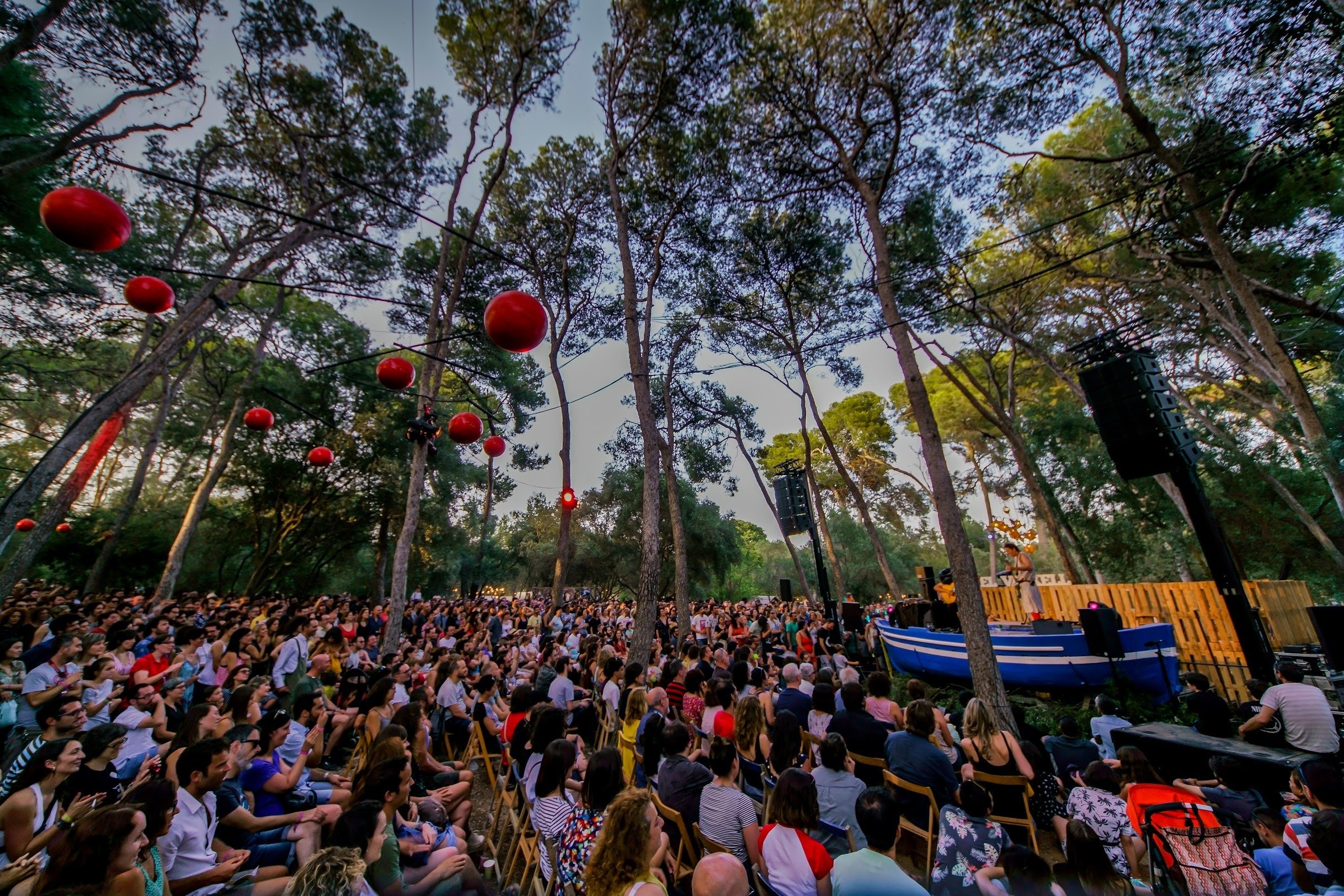 A large crowd of people sitting amidst tall trees, watching a performer at Vida Festival in Spain.